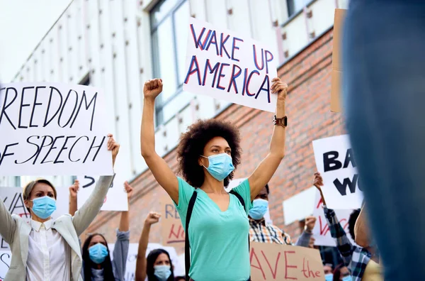 Low angle view of crowd of people wearing protective face mask while holding signs and banners on anti-lockdown protest. Focus is on black woman holding \'wake up America\' sign.