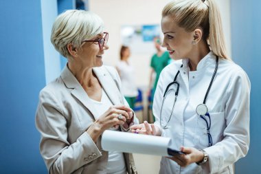 Happy mature woman and her doctor communicating while going through paperwork in a hospital hallway. 