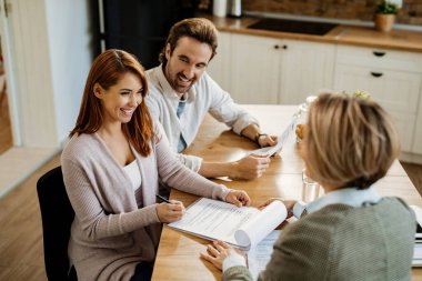 Young happy woman and her husband communicating with insurance agent and signing a document during a meeting. 