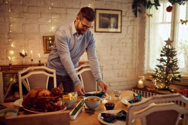 Smiling man making preparations for family dinner on Thanksgiving in dining room.