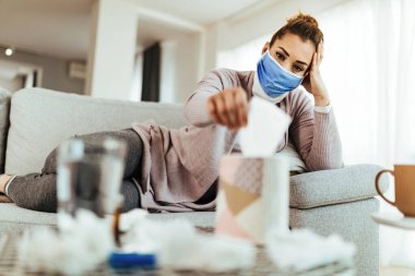 Pensive woman with face mask using paper tissue while relaxing on the sofa in the living room. 
