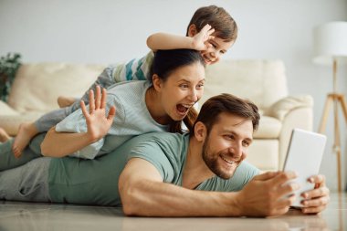 Happy family having fun while making human pyramid and taking selfie with digital tablet at home. 