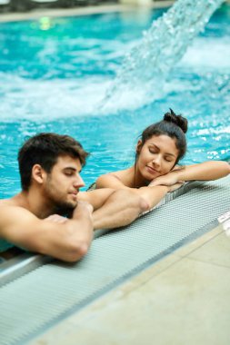 Relaxed couple leaning at the edge of a swimming pool and enjoying in the water with eyes closed at the spa. 