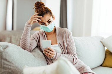 Distraught woman wearing protective mask while sitting on the sofa and text messaging on mobile phone at home. 