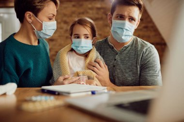Caring parents and their ill daughter wearing protective face masks while using computer at home.