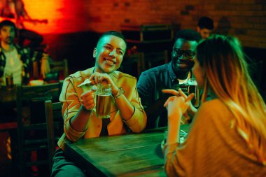 Young happy people drinking beer and communicating during their night out in a bar. Focus is on African American woman.