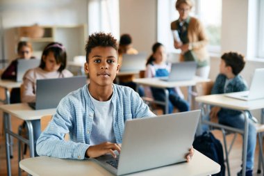 African American schoolboy e-learning on laptop in the classroom and looking at camera.