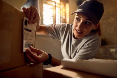 Young delivery woman preparing packages for shipment and attaching data label on carboard box in the office. 
