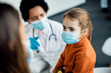 Small girl with face mask talking to her mother and doctor in a waiting room at the hospital. 