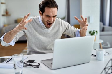 Young stressed freelance worker shouting while working on laptop at home. 