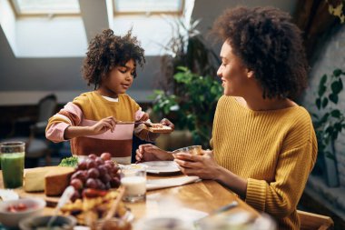 Little black girl spreading jam on toasted break while eating breakfast with mother in dining room.