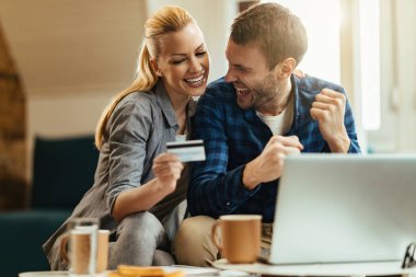 Happy couple having fun and celebrating while using credit card for e-banking at home.  clipart