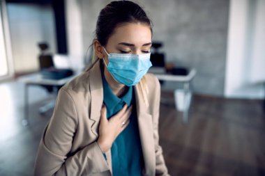 Distraught businesswoman holding her chest in pain while wearing face mask and working in the office during coronavirus pandemic. 