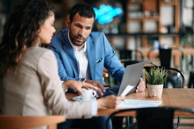 Young business coworkers analyzing paperwork on coffee break in a cafe. Focus is on businessman with laptop. 