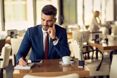 Smiling businessman writing in notebook while relaxing on coffee break in a cafe. 
