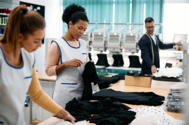 Young black tailor and her colleagues working with textile at clothing factory.