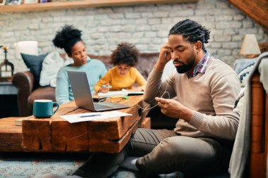 Evde dizüstü bilgisayar üzerinde çalışırken kulaklık kullanan Afro-Amerikalı bir adam. Arka planda karısı ve kızı var.. 