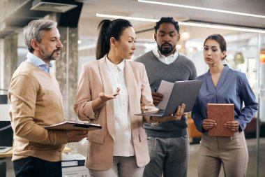 Multi-ethnic group of business coworkers using laptop during team meeting at corporate office. Focus is on Asian businesswoman. 