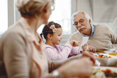 Happy girl showing something on mobile phone to her grandfather during a meal at dining table. 