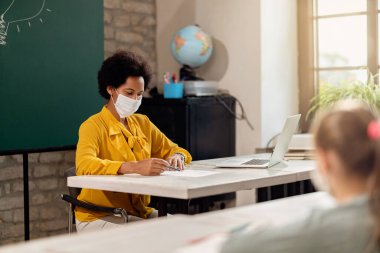 Black elementary school teacher with face mask reviewing students tests in the classroom. 