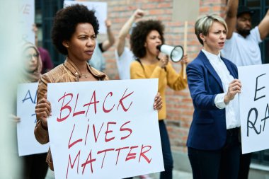 Crowd of people protesting for human rights on city streets. Focus is on African American woman with Black Lives Matter placard.