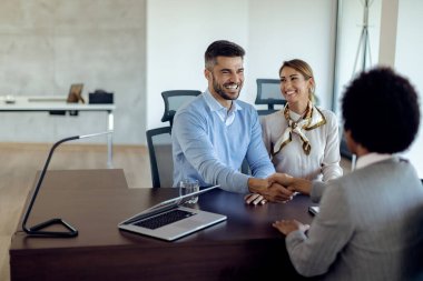 Young happy couple came to an agreement with real estate agent in the office. Focus is on man handshaking with the agent. 