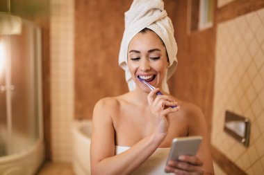 Young happy woman toothbrushing while text messaging on mobile phone in the bathroom.