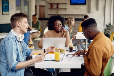 Happy African American student using laptop and talking to her friends at university cafeteria. 