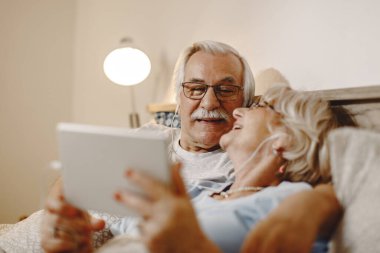 Happy senior man and his wife having fun while using digital tablet and relaxing in bed. 