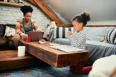 African American girl coloring at the table while her mother is working on laptop  at home.