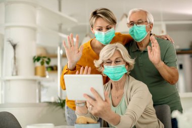 Senior couple and their adult daughter wearing protective face masks and greeting someone while making video call at home during coronavirus pandemic. 