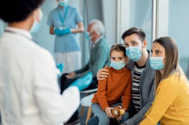 Young parents and their daughter communicating with a doctor while wearing face masks in waiting room at the hospital. 