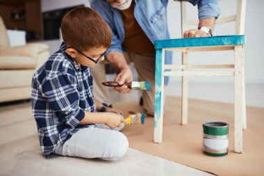 Small boy repairing furniture with grandfather and painting wooden chair in blue.