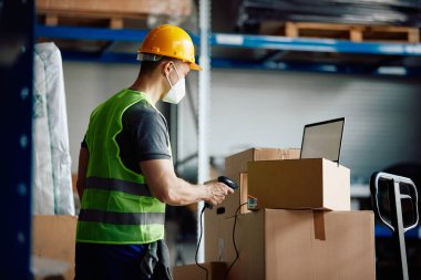 Warehouse worker with face mask using laptop and scanning packages at storage compartment.