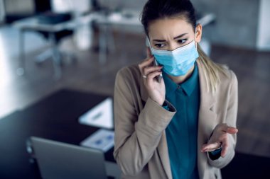 Displeased businesswoman making a phone call while working in the office during coronavirus pandemic. 