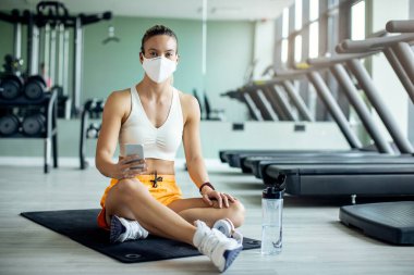 Athletic woman with face mask using smart phone while relaxing on the floor after working out in a gym during coronavirus epidemic.