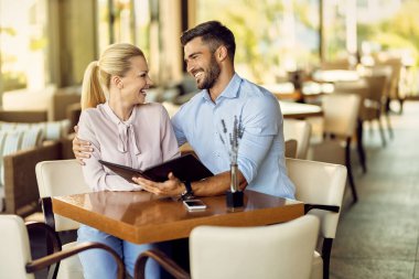 Happy man embracing his girlfriend while choosing something from a menu with her in a restaurant. 