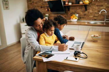 African American mother writing notes and working at home while children are sitting in her lap. 