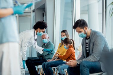 African American pediatrician communicating with a little girl in waiting room at medical clinic. They are wearing face masks due to COVID-19 pandemic.