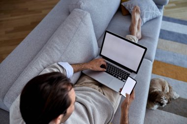 High angle view of a man relaxing on the sofa and using smart phone and laptop. Copy space. 