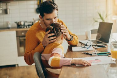 Affectionate father kissing his small daughter who is drawing on the paper while he works on computer at home. 