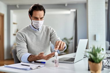 Young businessman with protective face mask using hand sanitizer while working on a computer at home. 