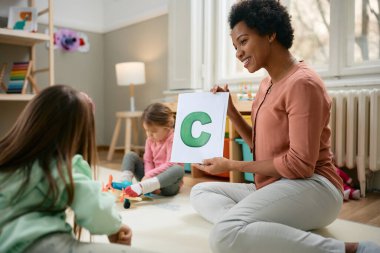 Happy African American teacher holding letter C while teaching kids the alphabet at kindergarten. clipart