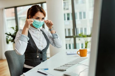 Young businesswoman using computer and putting on protective mask on her face in the office.