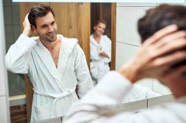 Reflection in a mirror of happy man in bathrobe in the bathroom. His wife is in the background. 