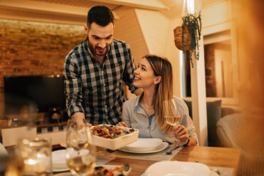 Young smiling man serving dinner to his girlfriend in dining room. 