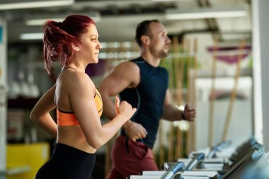 Athletic couple jogging on treadmill while exercising at health club. Focus is on woman. 