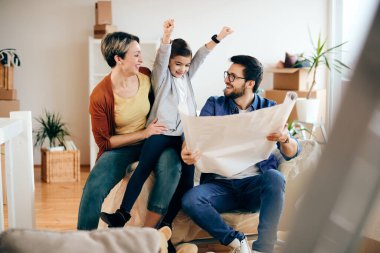Happy little girl with raised arms having fun while analyzing with her parents blueprints of their new home. 