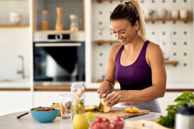 Young happy sportswoman slicing fruit while making smoothie in her kitchen. 