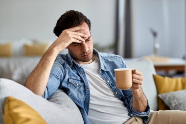 Young distraught man drinking coffee and thinking of something while sitting on the sofa. 